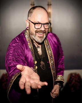 James Divine, Palm Reader, Mystic, Bringer of Joy. Image of smiling James, wearing purple velvet robes with his right hand outstretched towards you.